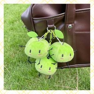 Puede incluir: Tres llaveros de peluche con forma de manzana verde, con brazos y piernas negros y caras sonrientes. Cada manzana tiene una hoja verde y está unida a un anillo de metal. Los llaveros cuelgan frente a una bolsa de cuero marrón sobre un fondo de hierba.