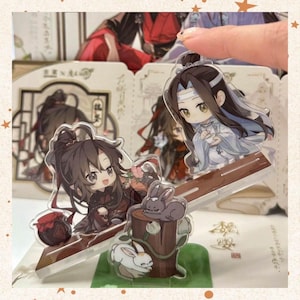 Peut inclure: Support en acrylique représentant deux personnages de style anime, l'un en robes noires et rouges, l'autre en blanc et bleu. Le support comprend une base en bois avec deux figurines de lapin. Le fond montre d'autres illustrations de personnages.