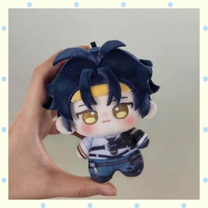 May include: A plush doll with dark blue hair, a yellow headband, and large, round eyes. The doll is wearing a white and blue outfit. The doll is held in a hand against a light background.