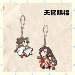May include: Two anime-style character charms with black lanyards. One character is in white robes, the other in red. The text "天官赐福" is above the charms. The background is a light beige colour with blue dots.
