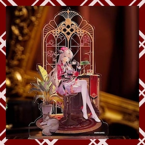 May include: Acrylic stand featuring an anime-style character seated in a decorative chair. The character wears a pink and red outfit, holding a teacup. A sleeping cat rests near a potted plant. The background includes a golden arch design.