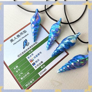 May include: Four iridescent blue seashell pendants on black cords. The pendants are arranged around a card with Chinese text and the English text "Mermaid's Pendant". The card has additional text and graphics.