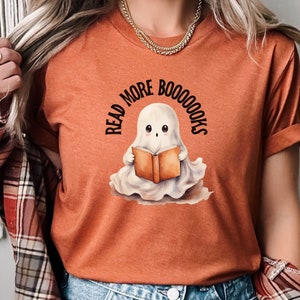 Op de afbeelding: Oranje T-shirt met een schattig spookje dat een boek leest. De tekst op het shirt luidt "Read More Booooks".