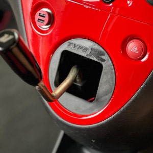 Può includere: Primo piano dell'interno di un'auto rossa con una leva del cambio nera e un emblema nero "TYPE R". L'auto ha un pulsante del freno di stazionamento rosso e una porta USB nera.