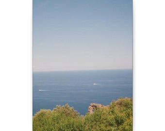 Könnte beinhalten: Leinwanddruck mit einer malerischen Küstenlandschaft. Das Bild zeigt eine weite Fläche des blauen Ozeans unter einem klaren, hellblauen Himmel. Grünes Laubwerk rahmt den unteren Bereich ein, mit zwei Booten auf dem Wasser.