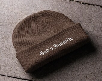 Puede incluir: Un gorro de punto marrón con la frase "God's Favorite" bordada en blanco. El gorro tiene un borde doblado y está hecho de un material texturizado. El gorro se muestra sobre una superficie gris.