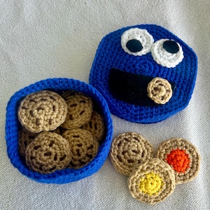May include: Blue crocheted cookie monster container with a removable lid. The container is filled with brown crocheted cookies. There are three additional cookies on the white surface next to the container. One cookie has a yellow center, one has an orange center, and one has a brown center.