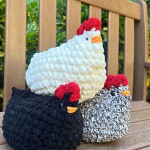 May include: Three crocheted chicken plushies sitting on a wooden bench. The chickens are white, black, and gray with red combs and orange beaks.
