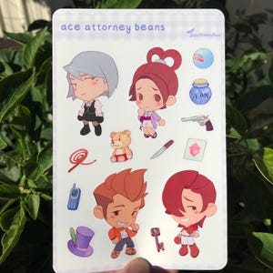 Peut inclure: Une feuille d'autocollants représentant des personnages de la série de jeux vidéo Ace Attorney. Les autocollants sont colorés et présentent divers personnages, dont un avocat, un détective et un ours en peluche. Le texte "ace attorney beans" est imprimé en haut de la feuille.