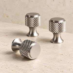 May include: Three silver-toned metal cabinet knobs with a cylindrical shape. Each knob features a textured, diamond-patterned knurled grip around the center and a smooth, flat circular top surface, supported by a tapered base that flares outward.