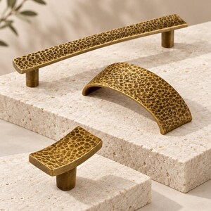 May include: Three brass cabinet hardware pieces with a hammered texture. The set includes one long bar pull, one arched pull, and one small rectangular knob. Each piece features a dark, antique-style finish with a dimpled surface pattern.