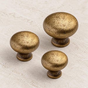 May include: Three round cabinet knobs in varying sizes, each featuring a textured, antique brass finish with a mottled gold and brown appearance. The knobs have a smooth, domed top and a tapered base, showcasing a consistent metallic surface.