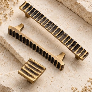 May include: A set of three brass cabinet hardware pieces including one square knob and two rectangular pulls. Each piece features a textured, dark-toned, ribbed surface with a metallic gold finish on the edges and sides.