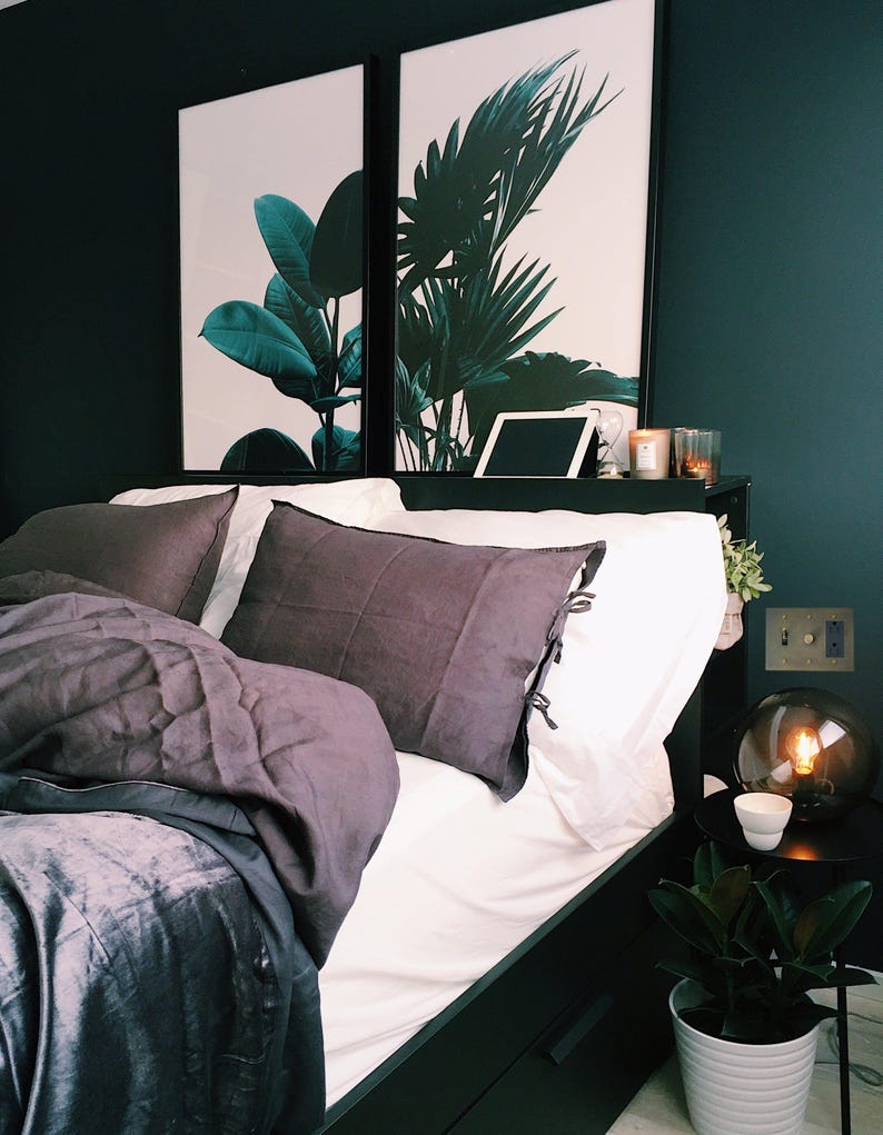 May include: A black bed frame with a white duvet and gray pillows. Two framed prints with green tropical leaves hang above the bed. A small table with a lamp and a potted plant sits next to the bed.