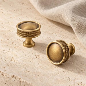 May include: Two round cabinet knobs with a brushed antique brass finish. Each knob features a smooth, slightly domed centre surrounded by a textured, cross-hatched band along the outer edge, supported by a tapered circular base.
