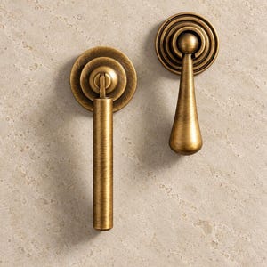 May include: Two antique brass cabinet pulls with a brushed finish. One pull features a long, cylindrical handle attached to a circular backplate, while the other has a teardrop-shaped handle mounted on a circular, tiered, concentric backplate.