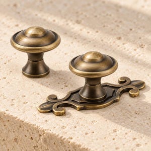 May include: Two antique brass cabinet knobs with a dark, brushed finish. One knob features a simple circular base, while the other is mounted on an elongated, decorative backplate with curved, scroll-like ends and a central pointed design.