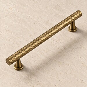 May include: A cylindrical cabinet pull handle featuring a textured, hammered finish in a muted antique brass tone. The hardware includes two conical mounting posts that connect the long, horizontal bar to a flat, circular base plate.