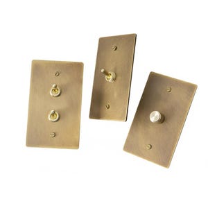 May include: Three antique brass light switch plates. One plate has two toggle switches, another has a single toggle switch, and the third has a single round button switch. All plates are rectangular with rounded corners.