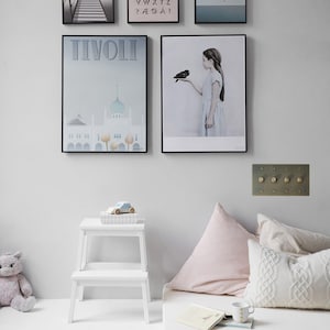 May include: A white wall with five framed posters, a white step stool, a white mattress with pink and white pillows, and a white rug. The posters feature a bridge, an alphabet, a girl with a bird, a building, and a girl with a cloud. The step stool has a toy car on top of it. The mattress has a book and a mug on top of it.