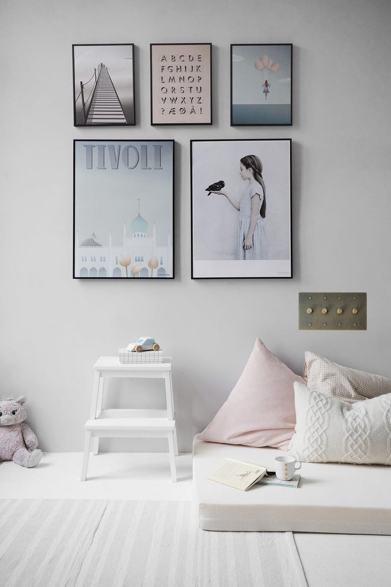 May include: A white wall with five framed posters, a white step stool, a white mattress with pink and white pillows, and a white rug. The posters feature a bridge, an alphabet, a girl with a bird, a building, and a girl with a cloud. The step stool has a toy car on top of it. The mattress has a book and a mug on top of it.