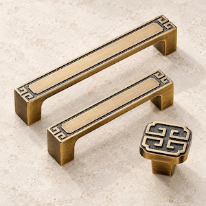 May include: Two rectangular cabinet pulls and one square cabinet knob in a brushed antique brass finish. Each piece features a dark, textured recessed border with a repeating geometric Greek key pattern at the corners and edges of the hardware.