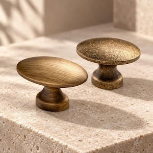 May include: Two oval-shaped cabinet knobs with a metallic finish. The knob in the foreground has a brushed brass texture, while the knob in the background features a darker, mottled antique bronze finish with a pedestal base for mounting.