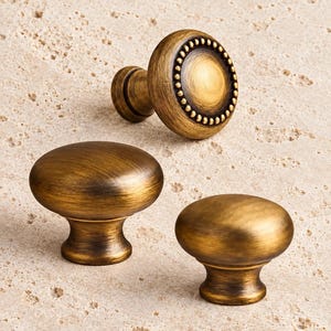 May include: Three cabinet knobs with a brushed antique brass finish. One knob features a circular beaded border around the center, while the other two knobs have a smooth, rounded dome shape with a tapered base and a dark, weathered patina.