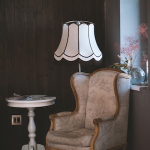 May include: A white lamp with a scalloped shade sits next to a beige armchair with floral upholstery. The chair has a high back and wooden legs. The lamp is on a wooden floor in a room with dark wood paneling.
