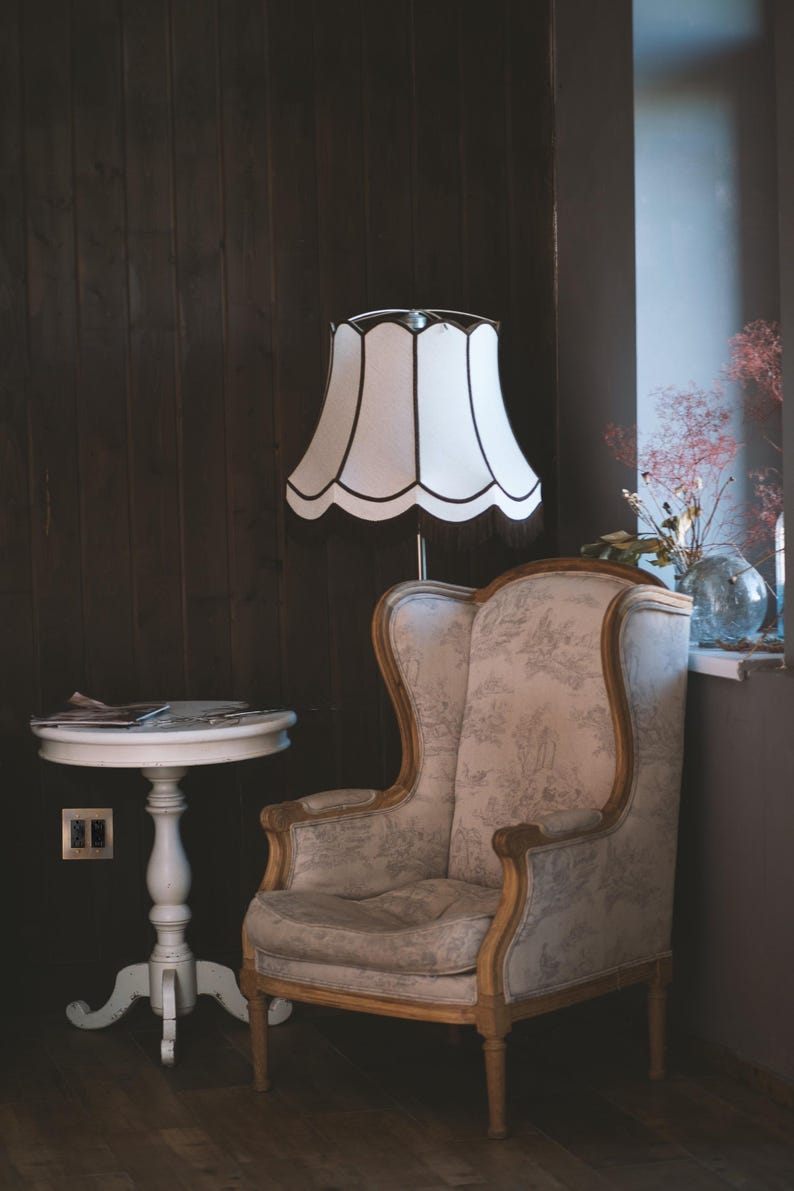 May include: A white lamp with a scalloped shade sits next to a beige armchair with floral upholstery. The chair has a high back and wooden legs. The lamp is on a wooden floor in a room with dark wood paneling.