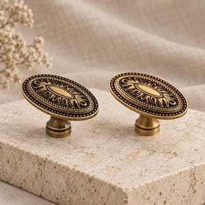 May include: Two oval-shaped cabinet knobs made of antique brass-toned metal. Each knob features a central smooth oval surrounded by a decorative sunburst pattern and a beaded outer rim, supported by a cylindrical base with a weathered finish.