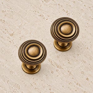 May include: Two round cabinet knobs with a brushed antique brass finish. Each knob features a series of concentric circular ridges that rise toward a smooth, rounded center point, supported by a short, cylindrical base pedestal.