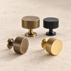 May include: Four cylindrical cabinet knobs featuring a vertical ribbed texture on the sides. Two knobs are finished in an antique brass tone, one is matte black, and one is a bright brushed gold, each with a central stem and circular base.