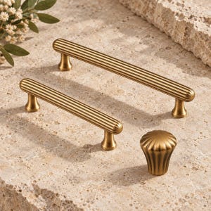 May include: Two brass-toned metal drawer pulls with a ribbed, cylindrical design and a single matching round, fluted cabinet knob. Each piece features a smooth, metallic finish with vertical grooves running along the length of the handles.
