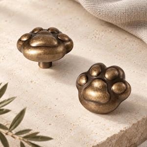 Puede incluir: Dos pomos para armarios con forma de huellas de animales, fabricados en metal de tono bronce oscuro con un acabado envejecido. Cada pomo presenta una almohadilla central y cuatro almohadillas de dedos más pequeñas en relieve.