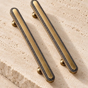May include: Two elongated brass cabinet pulls with a brushed metallic centre and a textured, dark-coloured border. Each handle features a smooth, rounded top surface and is supported by two small, circular mounting posts at the ends.