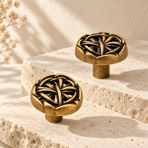 May include: Two round cabinet knobs made of antique brass with a dark, recessed finish. Each knob features a raised, symmetrical geometric pattern resembling a stylized butterfly or floral motif with a central vertical bar and radiating lines.