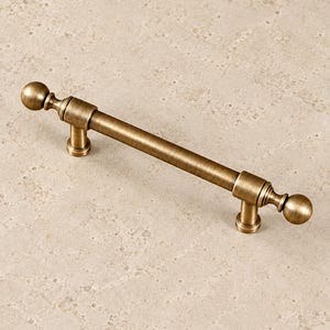 May include: A brushed brass cabinet pull featuring a long cylindrical handle with two mounting posts and decorative spherical finials at each end. The metal hardware has a warm, muted gold tone with a subtle horizontal brushed texture throughout.