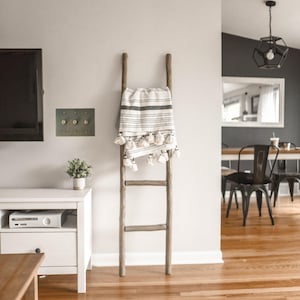 May include: A wooden ladder with a white and black striped throw blanket draped over it. The blanket has white tassels. The ladder is standing in a room with hardwood floors and a white rug.