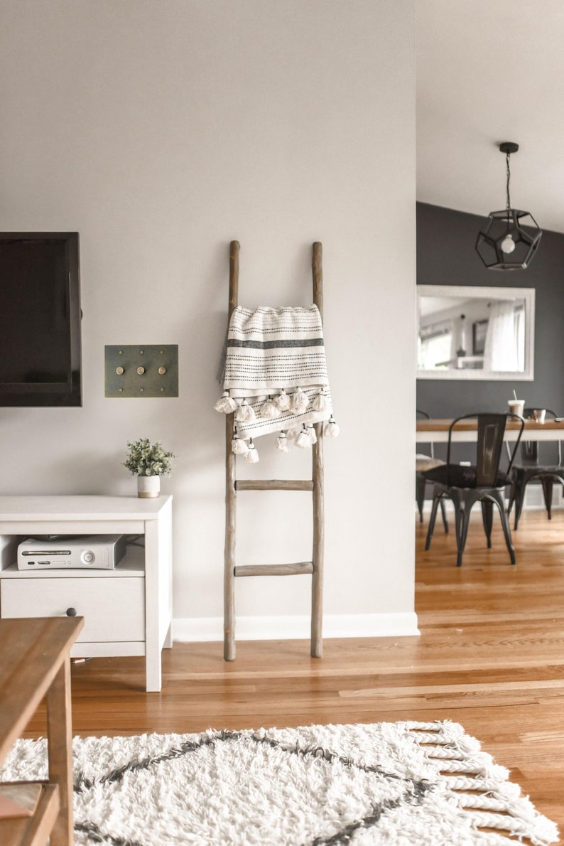 May include: A wooden ladder with a white and black striped throw blanket draped over it. The blanket has white tassels. The ladder is standing in a room with hardwood floors and a white rug.