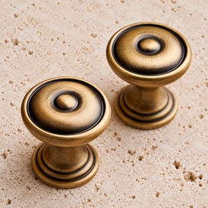 May include: Two round cabinet knobs with a brushed antique brass finish. Each knob features a circular, tiered base and a smooth, slightly domed top with a central raised button detail, showcasing dark, distressed accents in the grooves.
