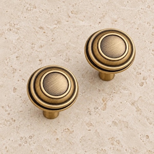 Peut inclure: Deux boutons de placard ronds avec une finition en laiton antique brossé. Chaque bouton présente un design circulaire concentrique à gradins avec un centre lisse légèrement bombé et un anneau extérieur surélevé, créant un aspect superposé.