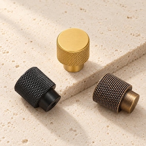 Könnte beinhalten: Drei zylindrische Schrankknöpfe mit einer texturierten, gerändelten Oberfläche im Rautenmuster. Ein Knopf ist in glänzendem gebürstetem Gold, einer in Mattschwarz und einer in Antikbronze gehalten, jeweils mit einer glatten, runden Oberseite.
