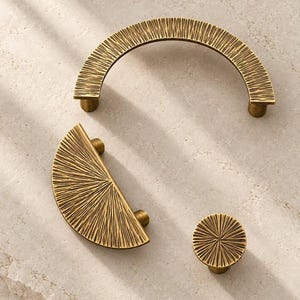 May include: A set of three brass-toned metal cabinet hardware pieces featuring a textured, sunburst-inspired pattern. The collection includes one large semi-circular arch handle, one smaller semi-circular pull, and one circular knob with a central point.