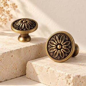 Könnte beinhalten: Zwei runde Schrankknöpfe aus antik messingfarbenem Metall. Jeder Knopf zeigt ein erhabenes, zentrales Blumenmotiv mit detaillierten, strukturierten Blütenblättern, die von einer kleinen, glatten kreisförmigen Mitte auf dunklem Grund ausgehen.