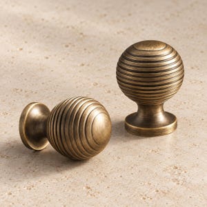 May include: Two spherical cabinet knobs with a brushed antique brass finish. Each knob features a series of horizontal, concentric circular grooves etched into the surface, creating a ribbed texture that wraps around the round head.