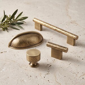 May include: A collection of four brass cabinet hardware pieces featuring a hammered texture. The set includes one long bar pull, one cup pull, one T-shaped knob, and one round knob, all finished in a muted, brushed metallic gold tone.