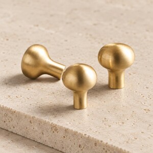 May include: Three mushroom-shaped cabinet knobs with a smooth, brushed gold finish. Each knob features a rounded top and a cylindrical base, designed for attachment to furniture or cabinetry. The metal surface has a soft, matte metallic sheen.
