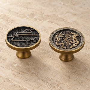 May include: Two round brass cabinet knobs with a brushed finish. One knob features a dark textured background with raised cloud motifs. The other knob displays a detailed relief of a dragon and a phoenix encircled by a geometric border pattern.