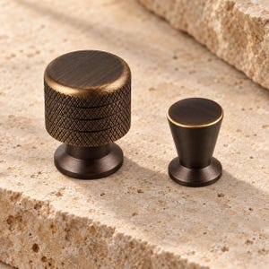 May include: Two dark bronze cabinet knobs with a metallic finish. The larger knob features a cylindrical shape with a textured, diamond-patterned knurled side. The smaller knob has a smooth, tapered, conical body with a flat circular top surface.